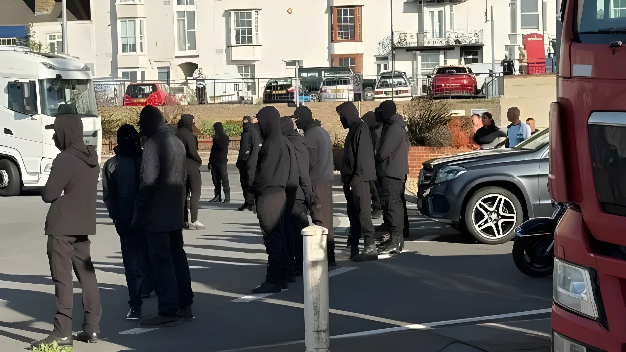 A row of masked men block a roundabout. Cars and lorries are stationary on the road.