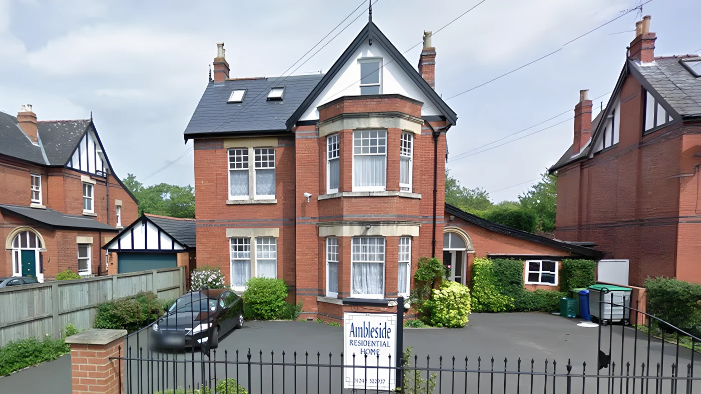 A screenshot from Google street view showing the care home. It is a large detached three-storey brick house with bay windows and a driveway outside the front. There are black railings around the property and a black car parked outside.