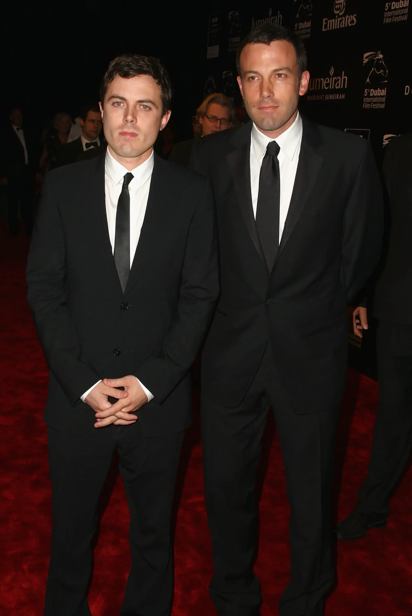 Casey Affleck and Ben Affleck in black suits on a red carpet.