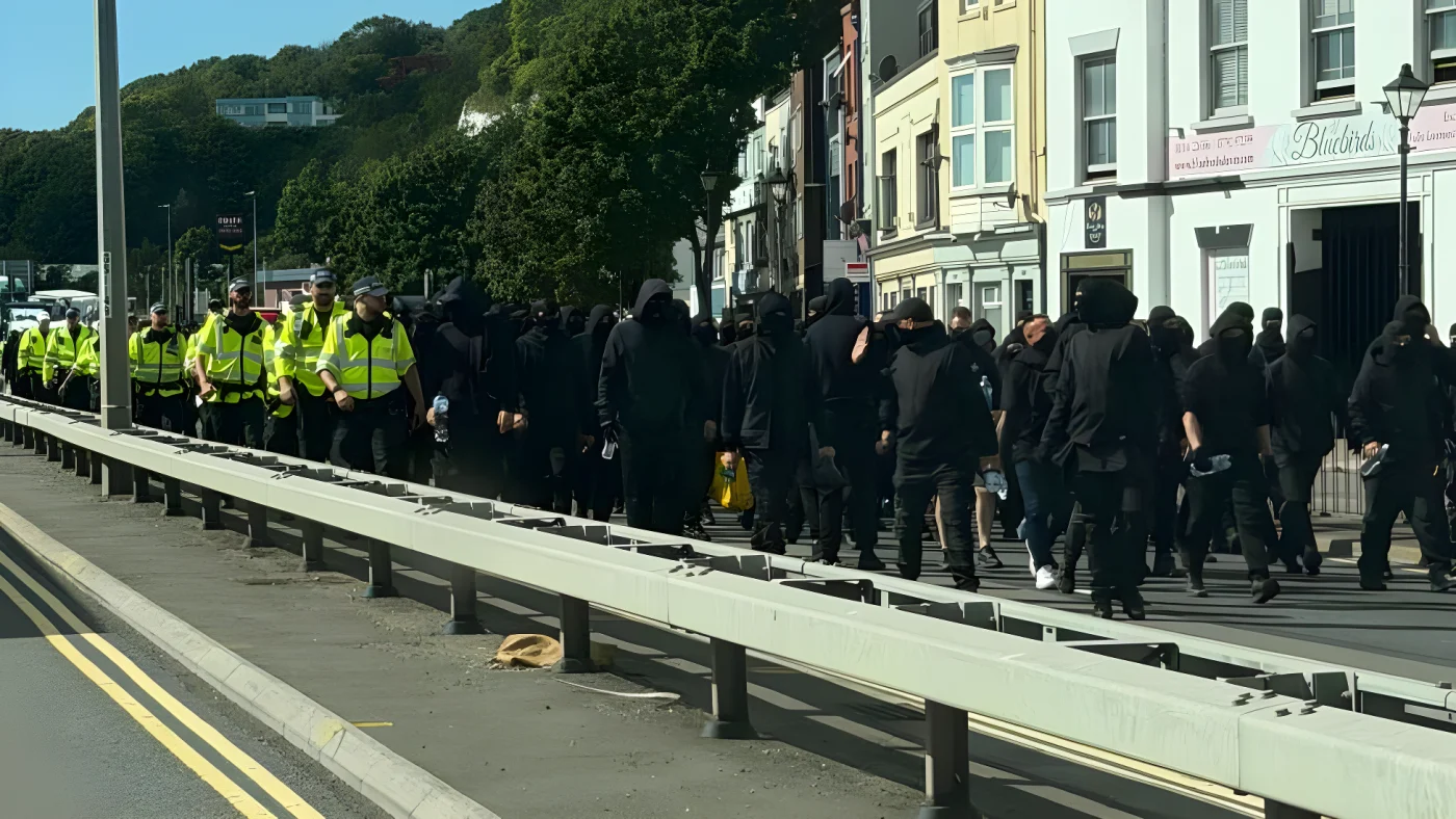 Dozens of men dressed in black and wearing face coverings, being escorted down a dual carriageway in Dover by uniformed police officers.