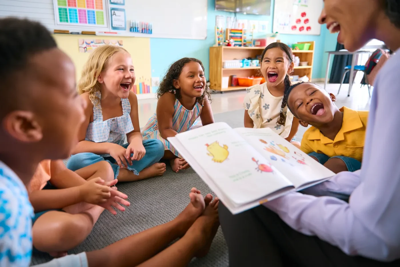 Female Primary Or Elementary School Teacher Reads Story To MultiCultural Class Seated In Classroom