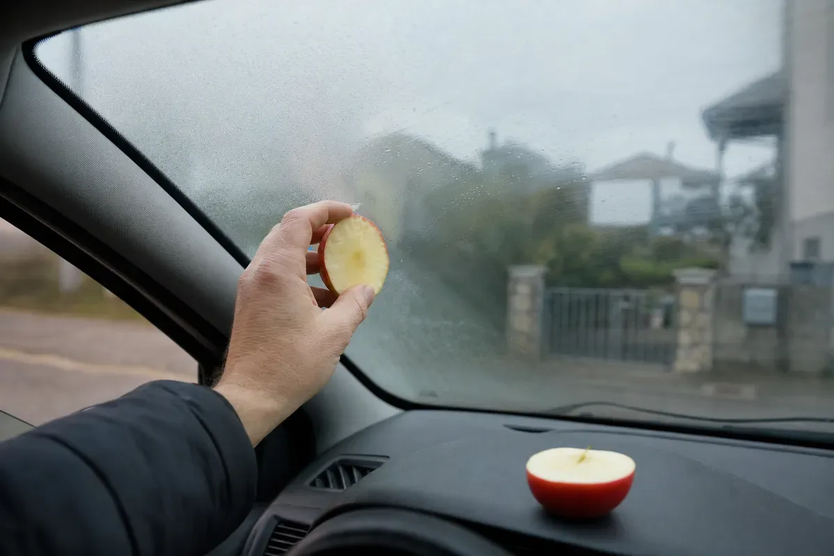 Mano che passa mezza mela sul parabrezza appannato dall’interno di un’auto, con l’altra metà sul cruscotto