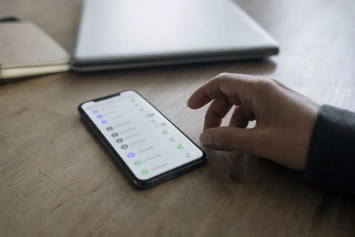 Smartphone su un tavolo con schermata impostazioni sfocata e una mano esitante vicino, laptop sullo sfondo