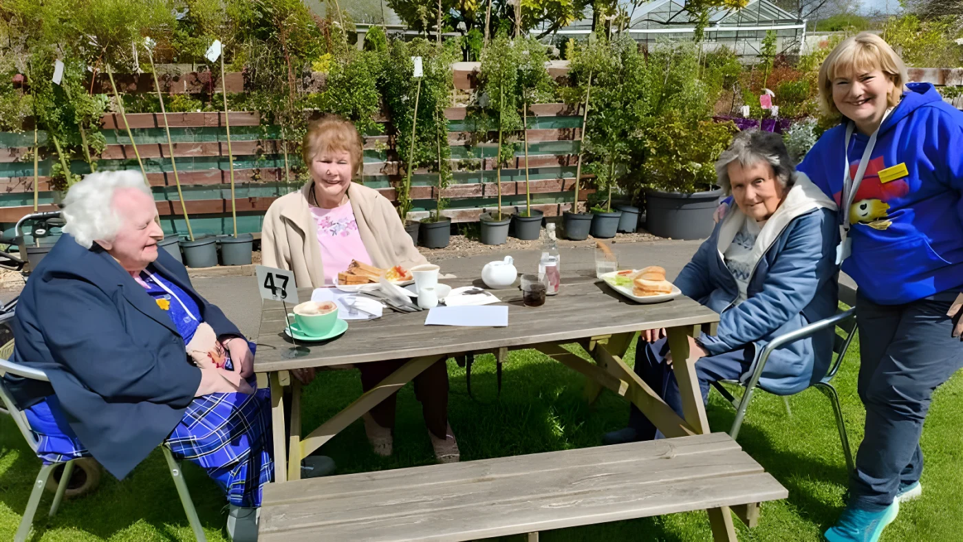 Three elderly women sit around a picnic bench on a lawn at a garden centre surrounded by trees for sale. A carer has her arm around one of the women and is wearing a blue Paddington Bear hoodie.