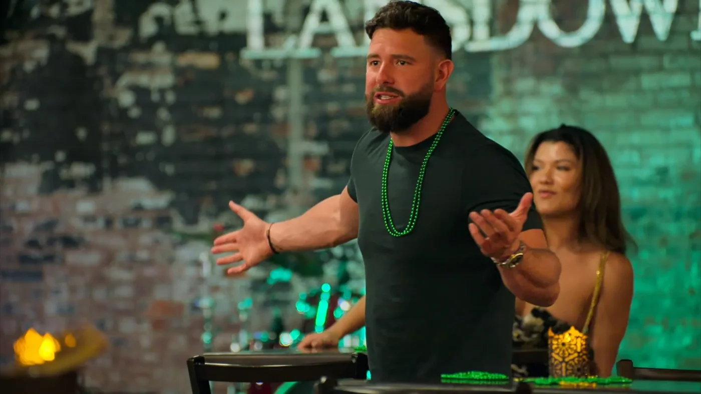 A bearded man wearing green beads speaks with his arms outstretched, with a woman sitting in the background.