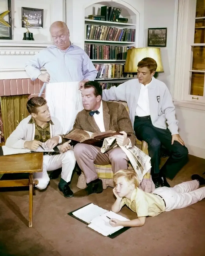 A black-and-white TV family gathers in a book-lined study: William Frawley stands holding a sheet, Fred MacMurray reads from a large book seated, Tim Considine leans over his shoulder, Don Grady writes at a desk, and young Stanley Livingston lies on the floor writing on a notepad.