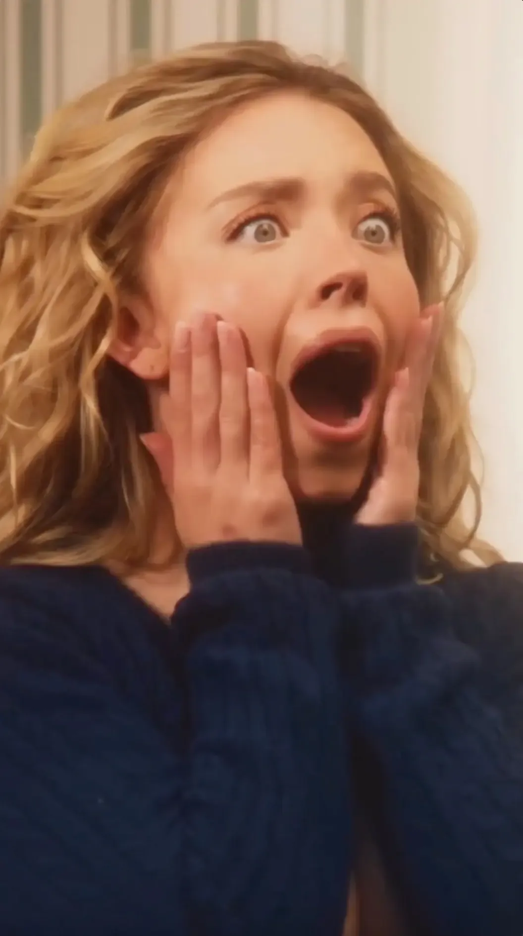 A blonde woman in a blue sweater gasping with her hands on her cheeks.
