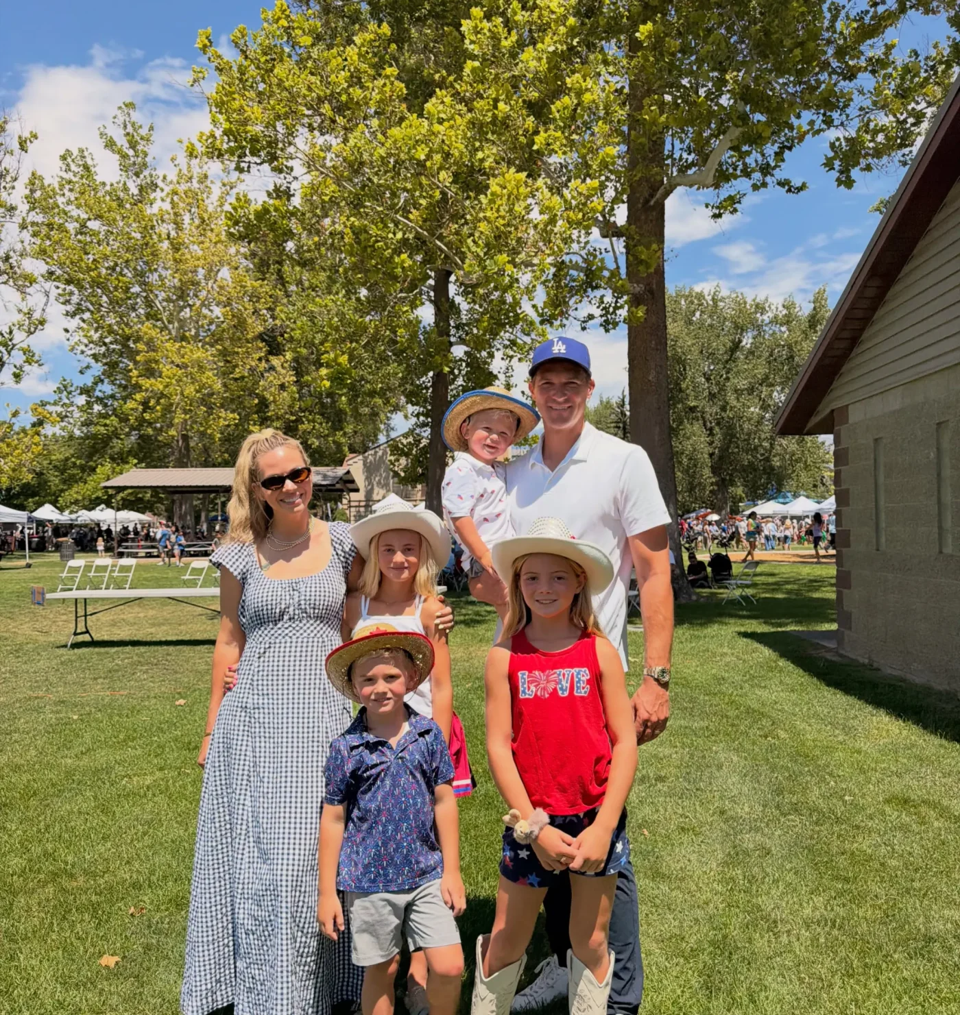 A family of six poses outside on a sunny day in a grassy park, with a mother, father, and four children wearing hats.