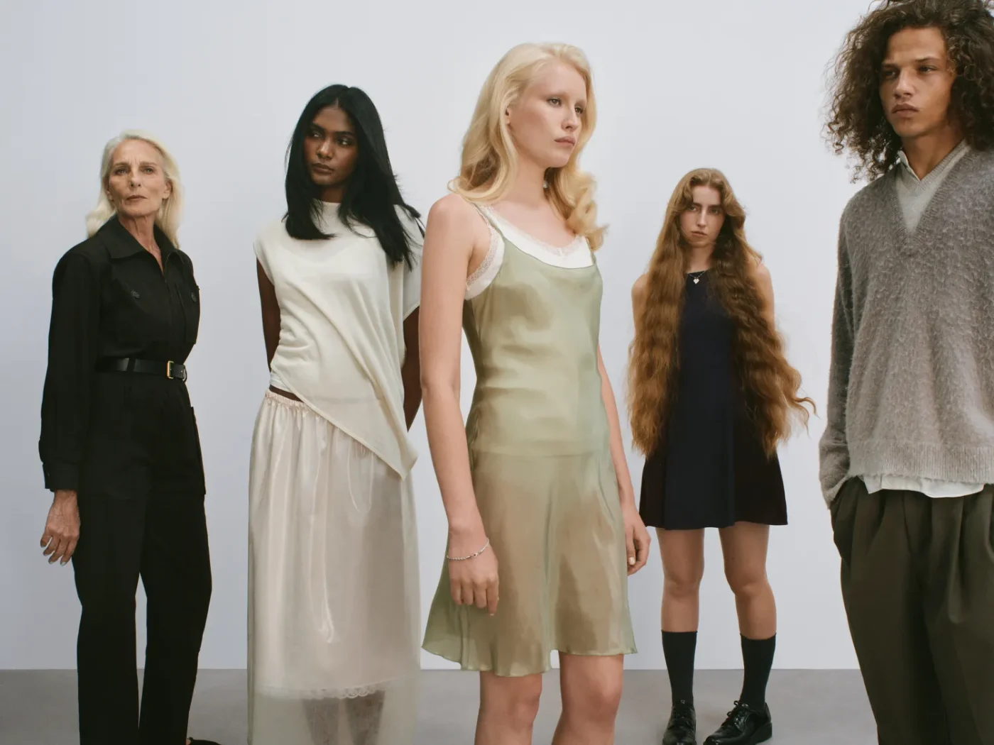 A group of people standing together wearing casual and formal attire with varied expressions and stances in a minimal setting