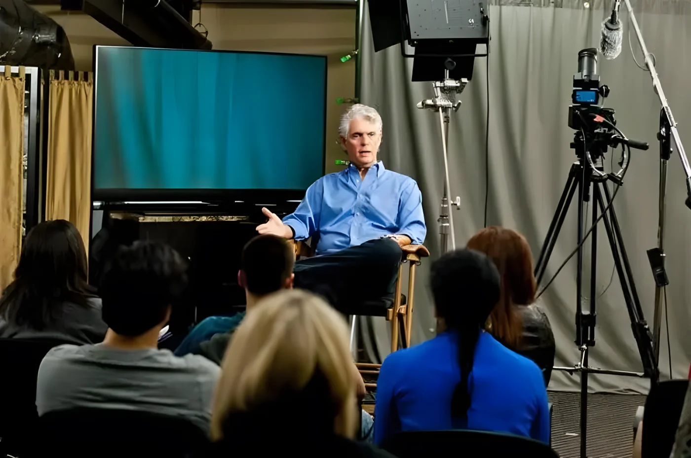 A man in a blue shirt speaks in front of a live audience in a television studio.