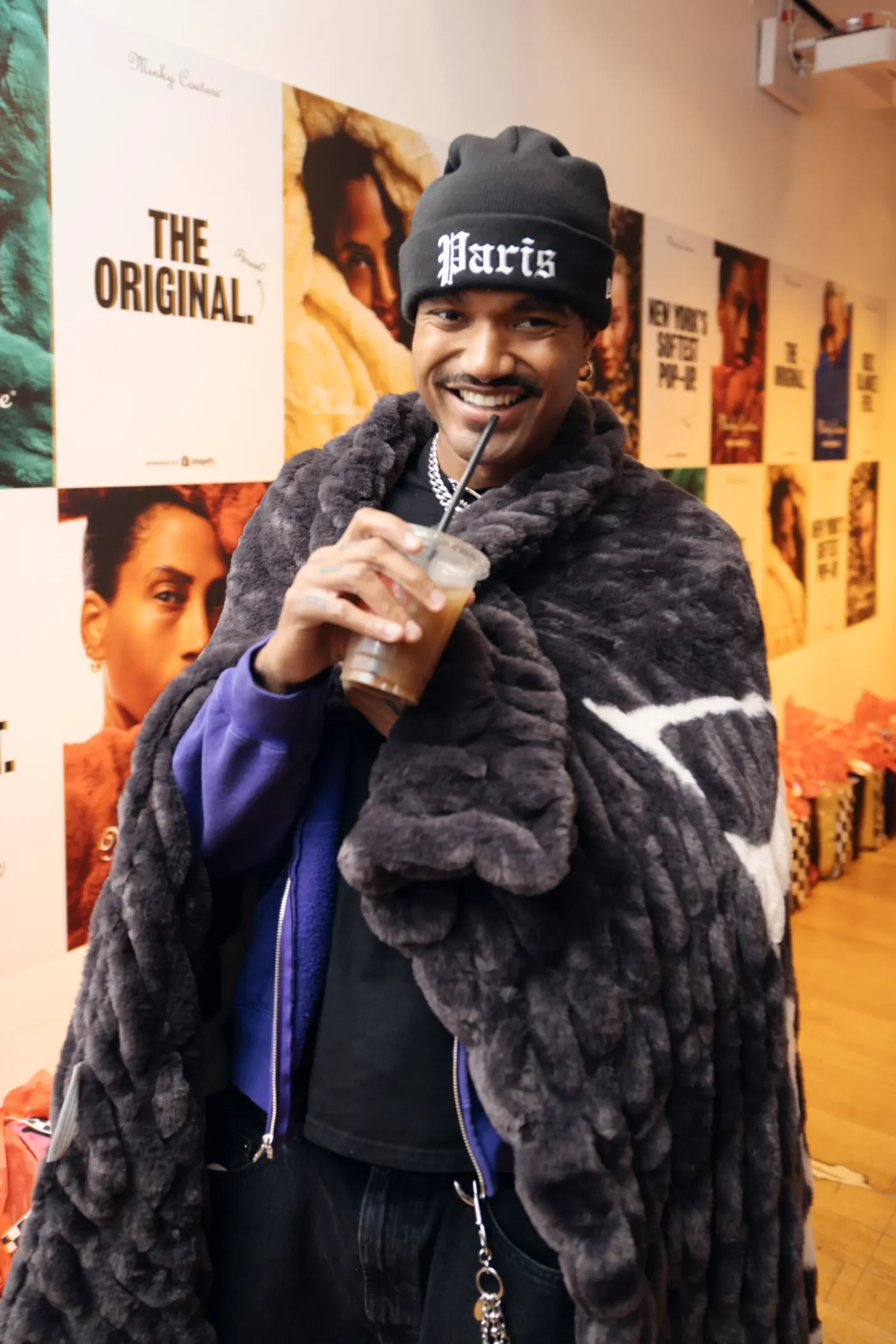 A man wearing a black beanie and a grey faux-fur blanket draped over his shoulders, smiling while holding an iced coffee in a plastic cup.