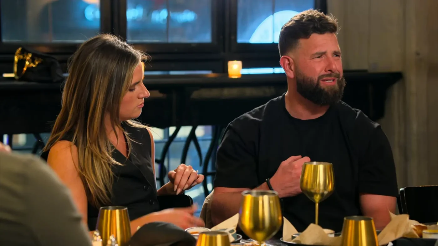 A man with a beard and a woman sit at a dimly lit restaurant table, speaking to each other.