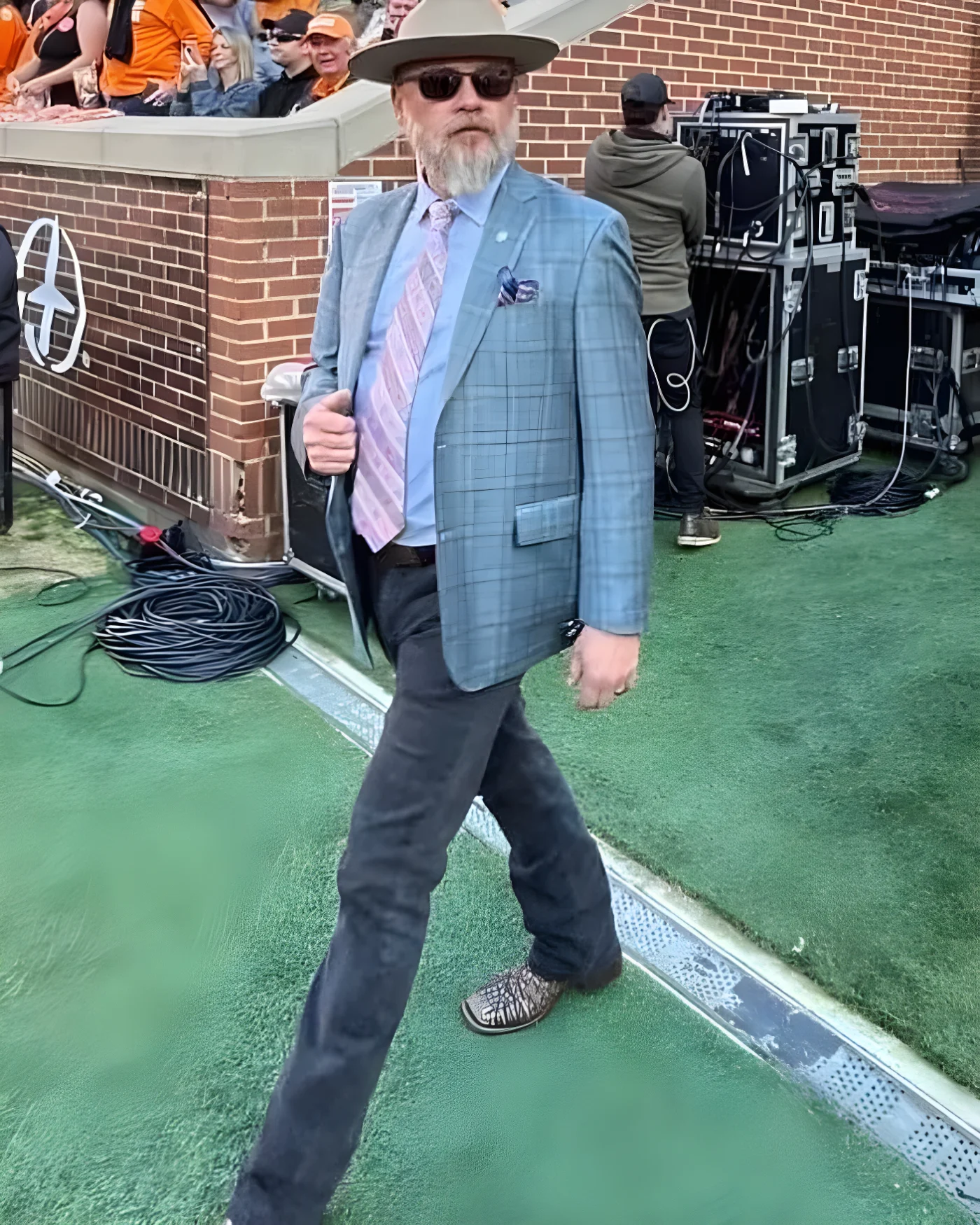 A man with a graying beard, sunglasses, and a fedora walks on a turf field, wearing a plaid sport coat, patterned tie, dark jeans, and boots.