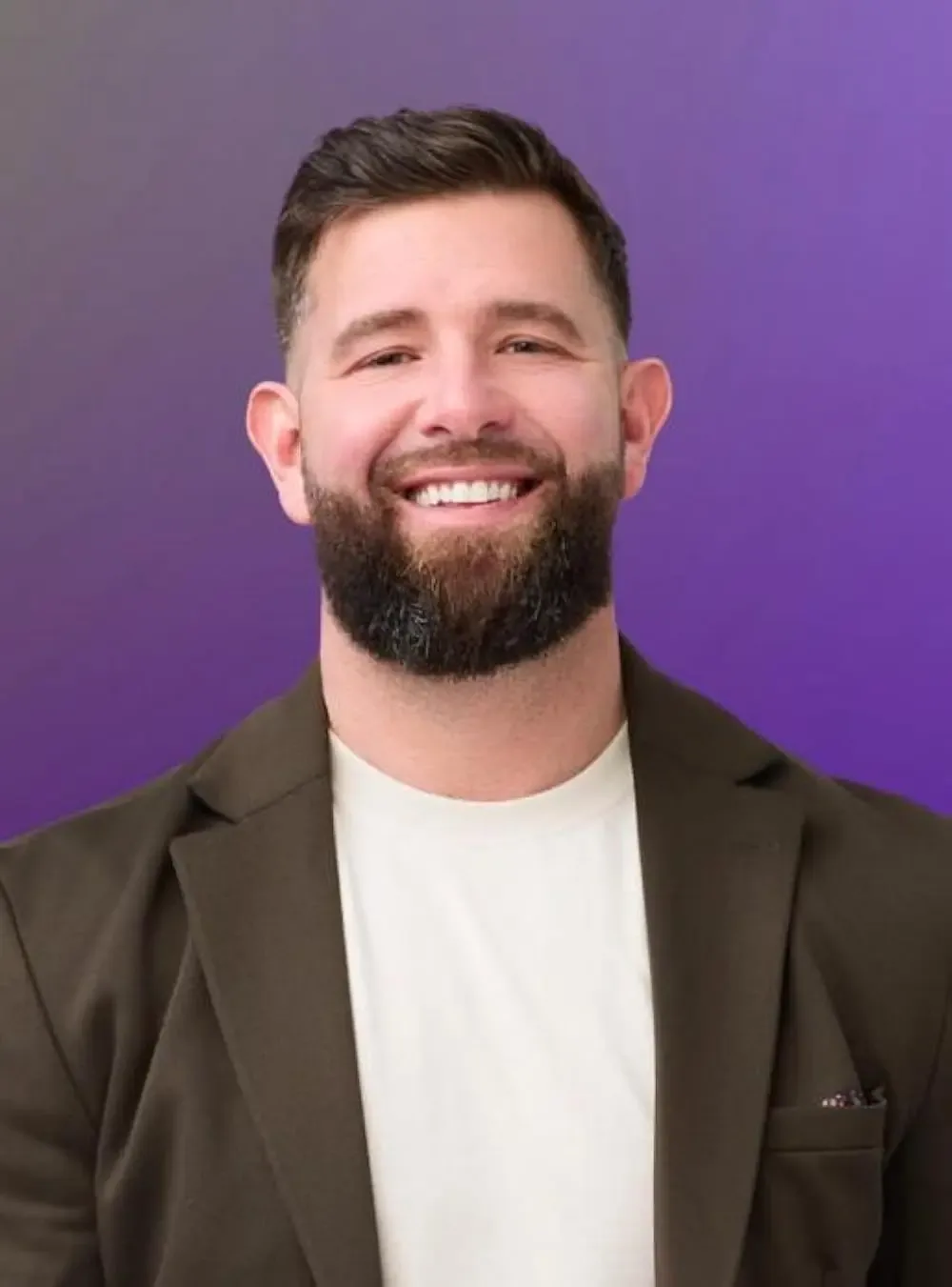 A man with short brown hair and a beard, wearing a white t-shirt and a dark green blazer, smiling against a purple background.