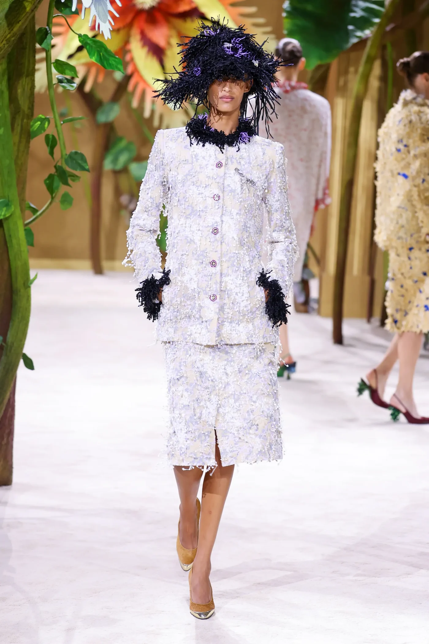 A model wearing a light-colored tweed jacket and skirt ensemble with dark, feathery accents walks the runway at Paris Fashion Week.