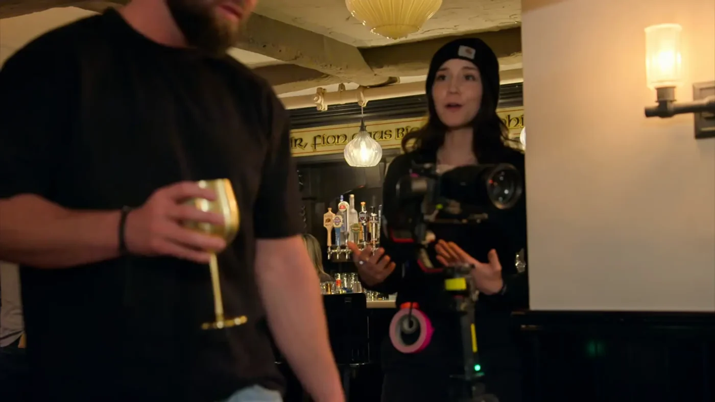 A person in a black shirt holds a golden goblet in a dimly lit bar, while another person operates a professional camera nearby.