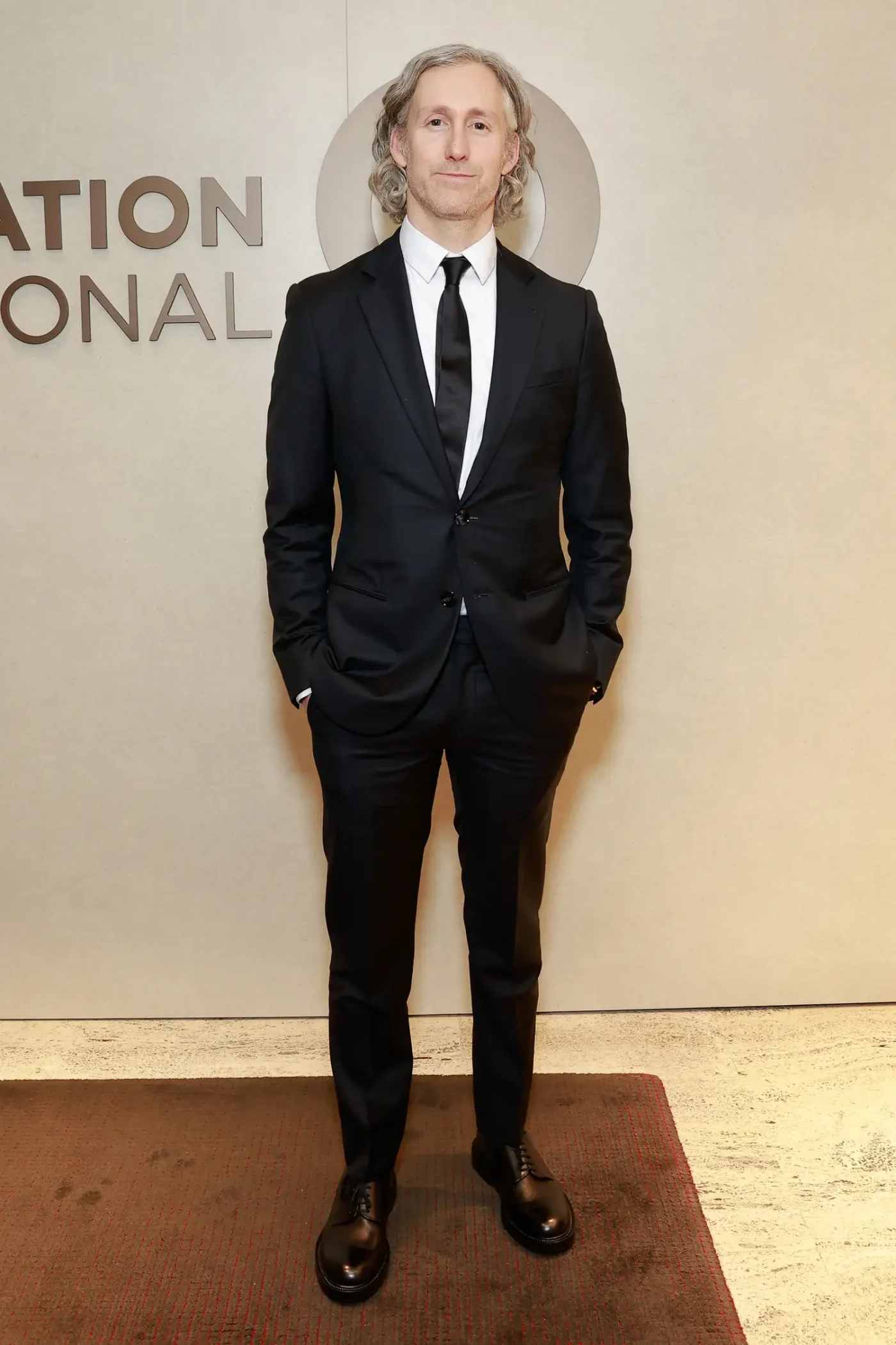 A person in formal attire stands indoors near a wall with partial text visible in the background
