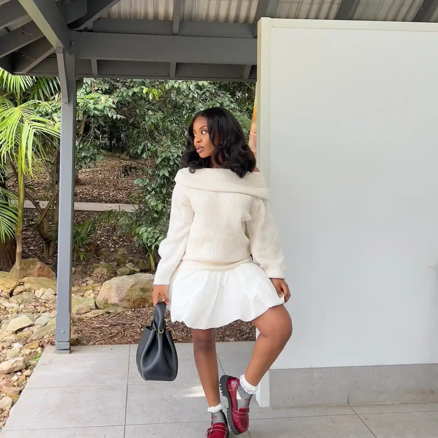 A person posing outdoors wearing a light outfit with a bag in hand