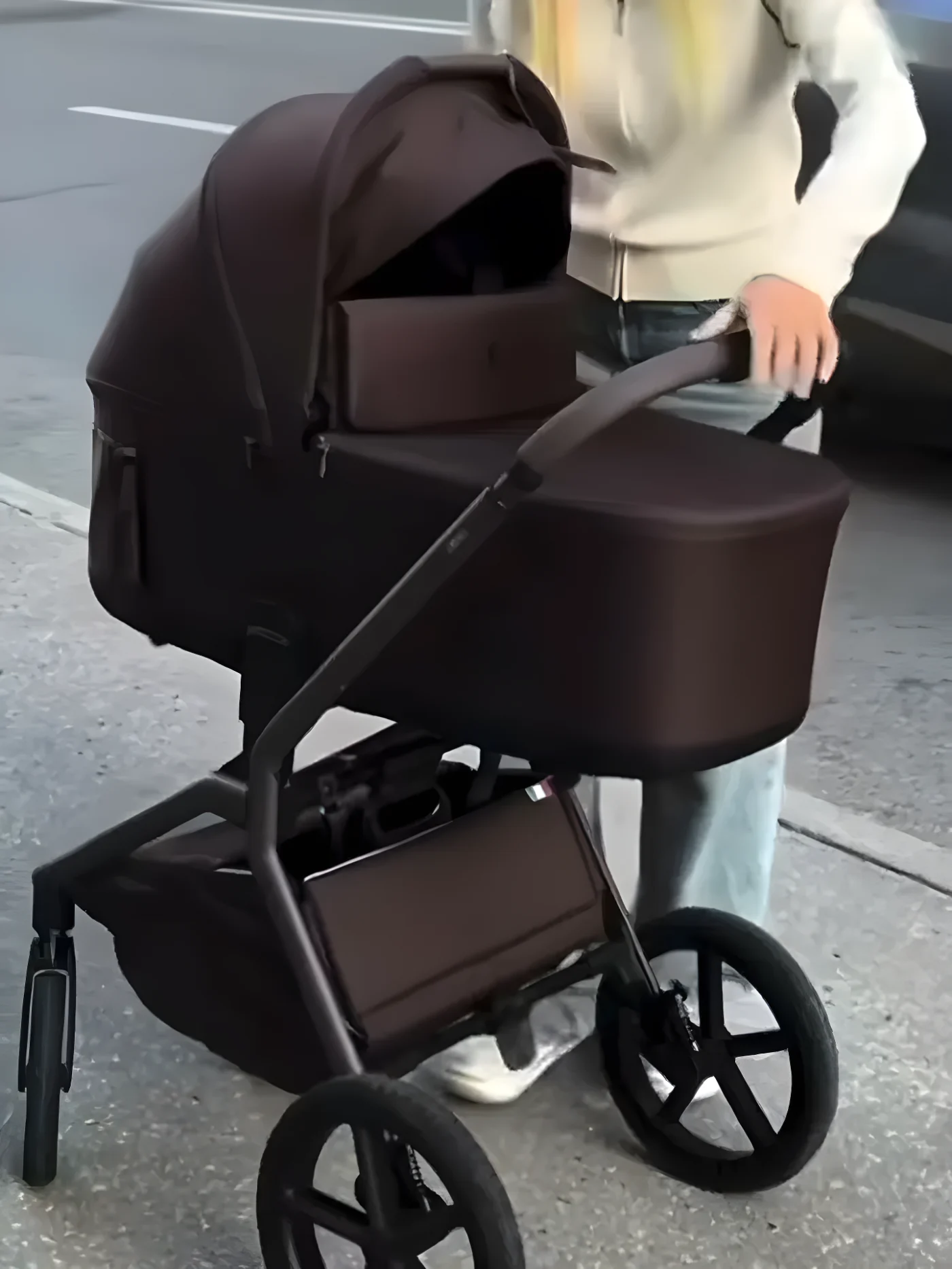 A person pushing a brown baby stroller containing a portable speaker on a sidewalk.