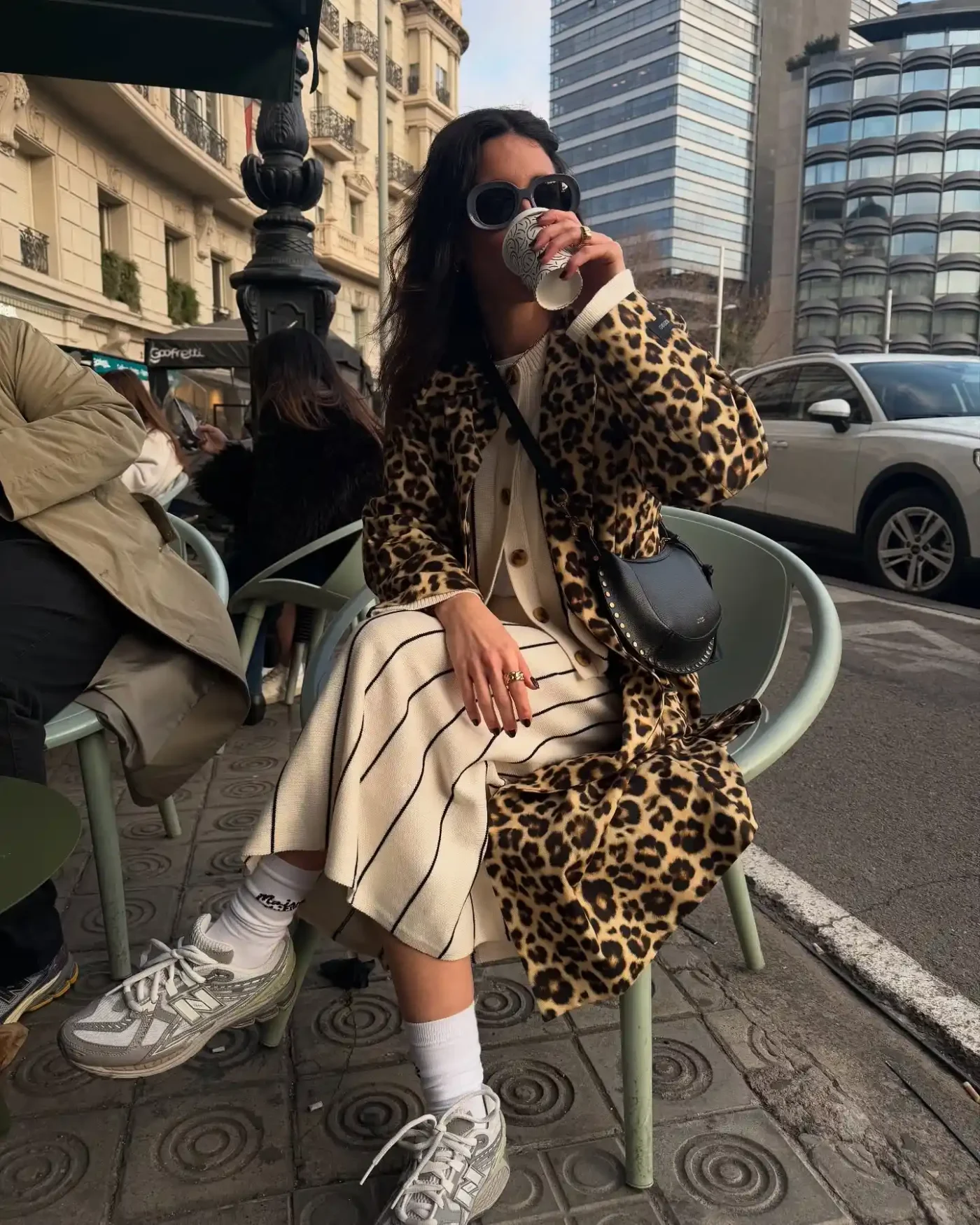 A person seated at an outdoor cafe drinks from a cup dressed in a patterned coat and casual attire
