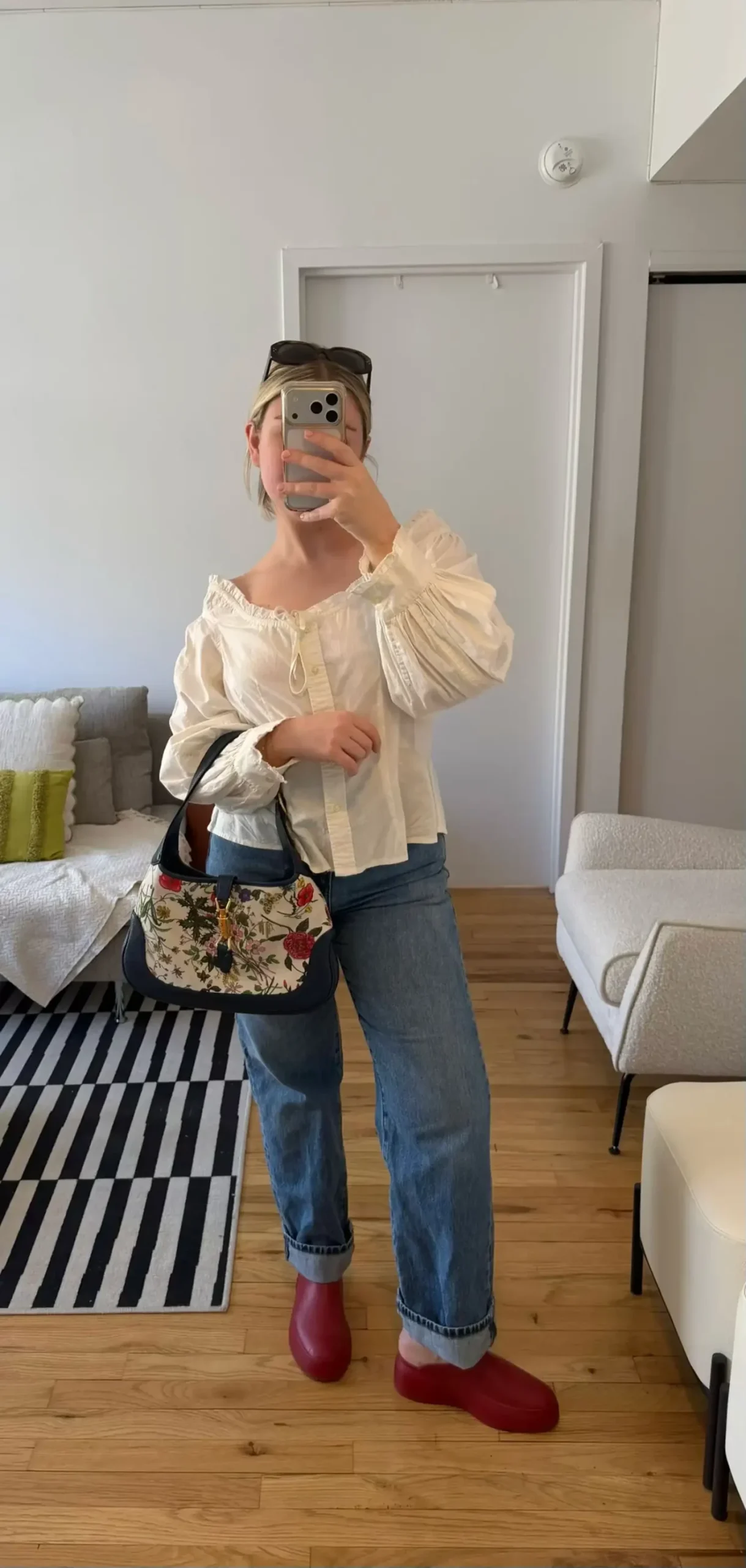 A person stands in a room taking a mirror selfie wearing relaxed clothing and holding a small patterned bag