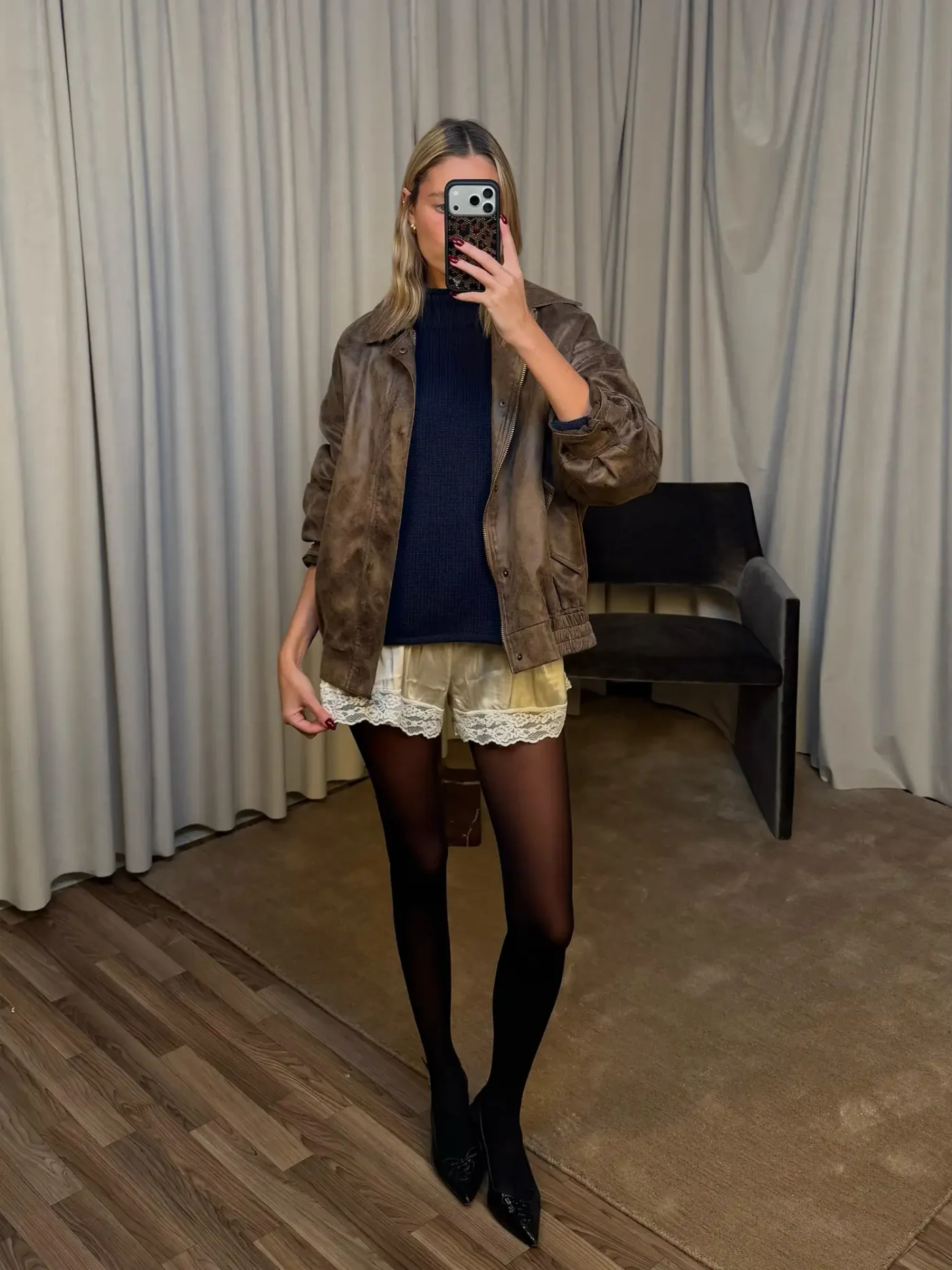 A person stands taking a mirror selfie indoors wearing a casual outfit with layered clothing and a neutral expression