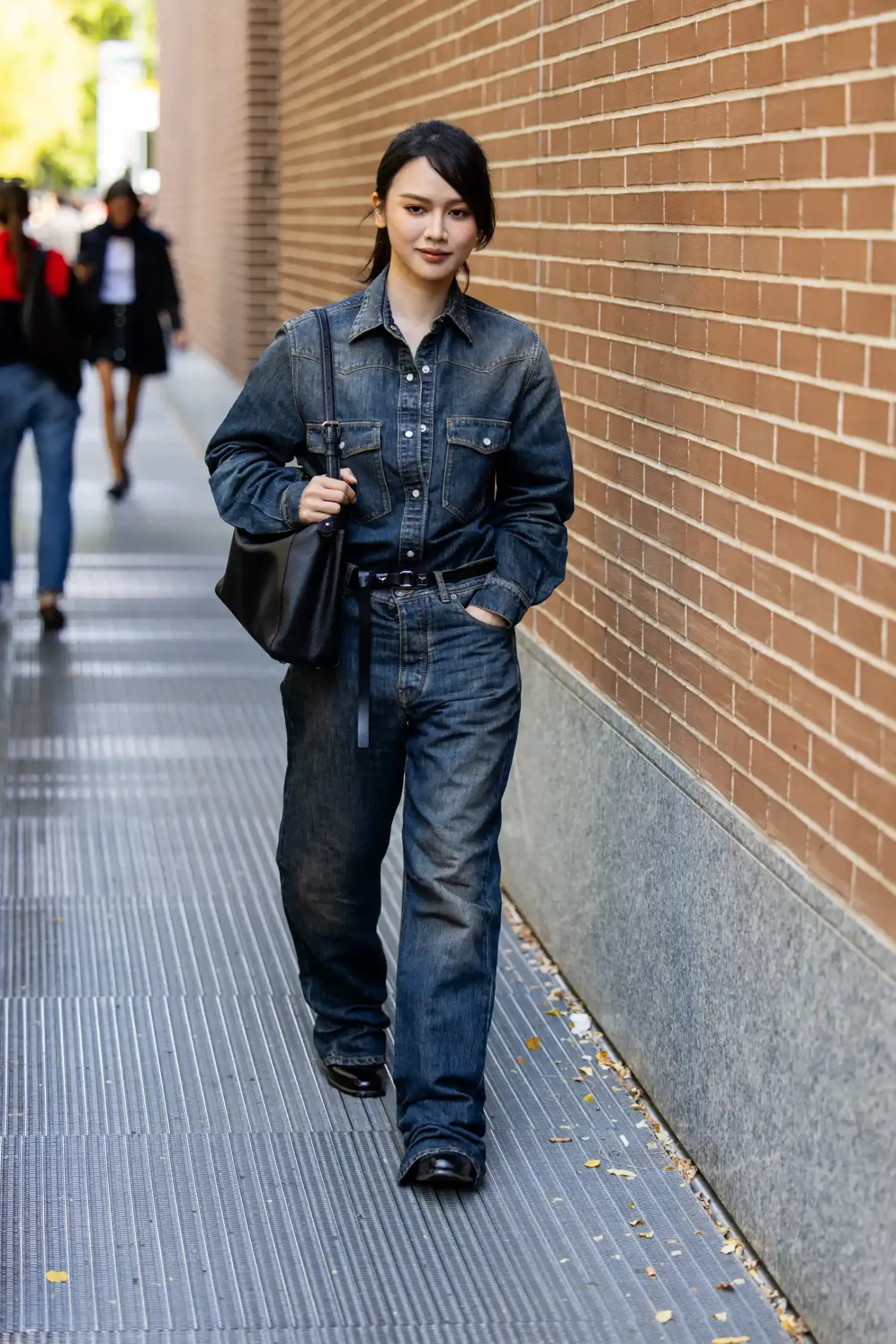 A person walking on a sidewalk beside a brick wall wearing denim clothing and holding a bag