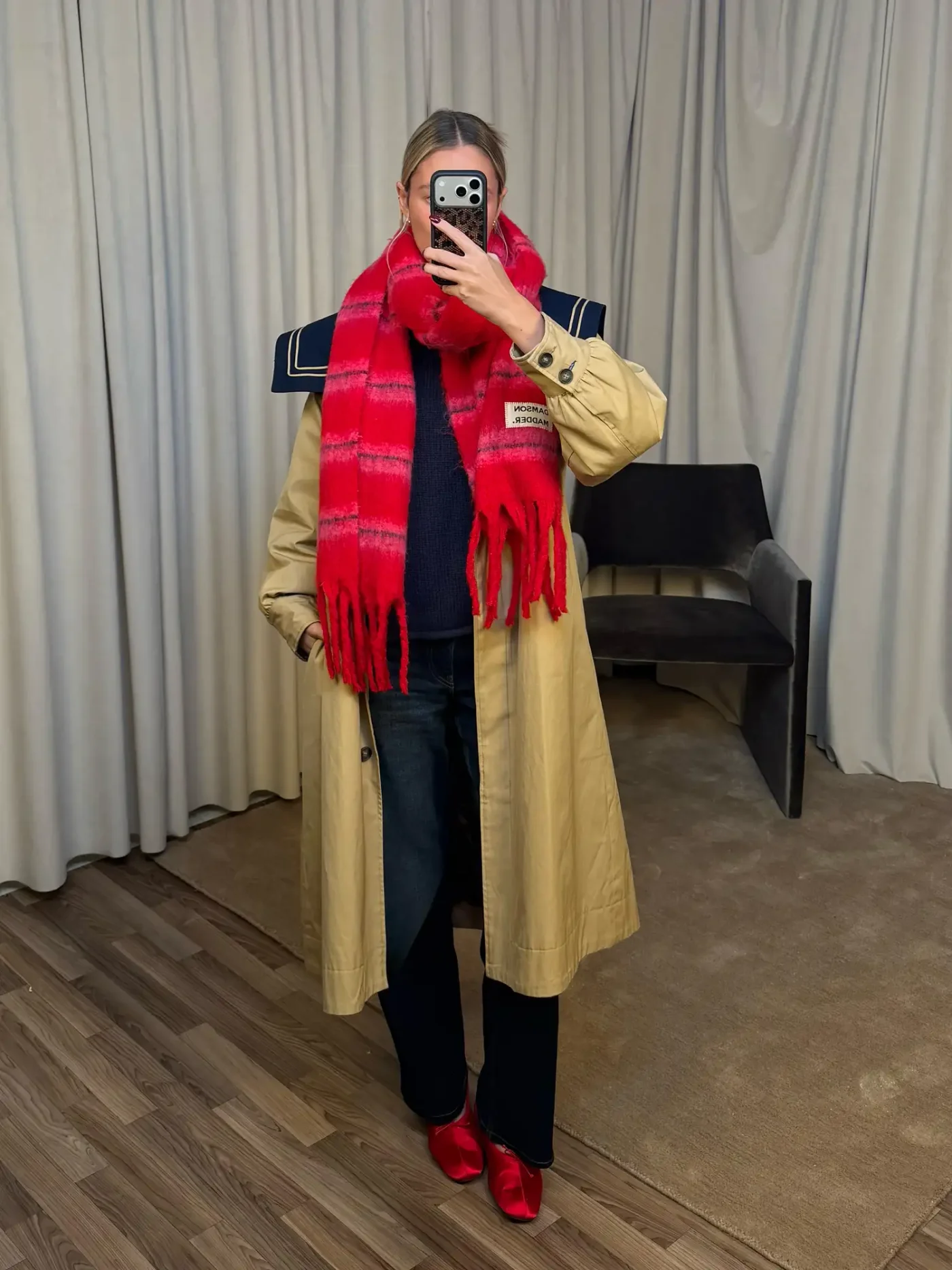 A person wearing a beige coat with a red scarf taking a selfie indoors while standing on a wooden floor