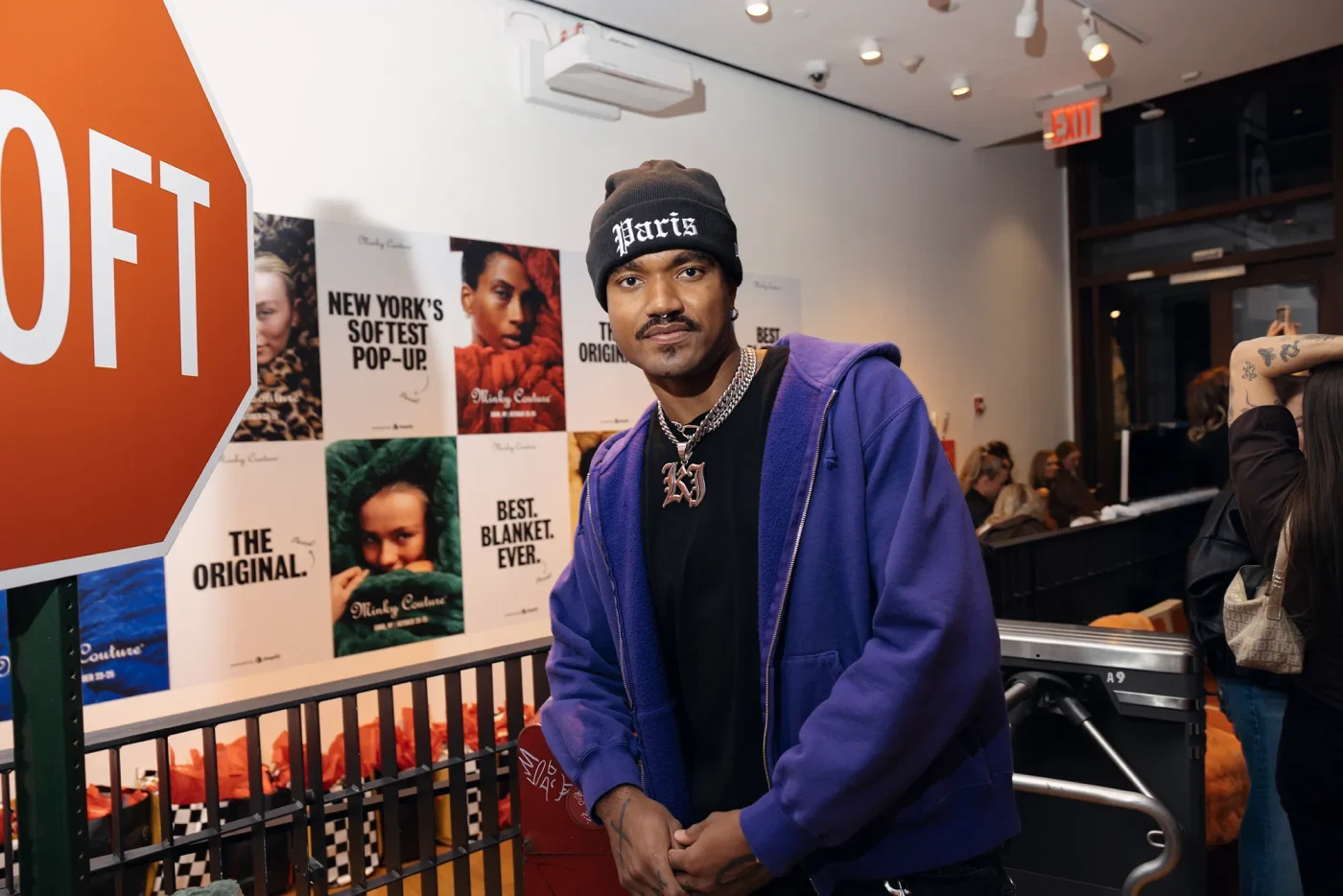 A person wearing a purple zip-up hoodie, black beanie, and silver chain necklace poses in an indoor setting with promotional posters on the wall behind them.