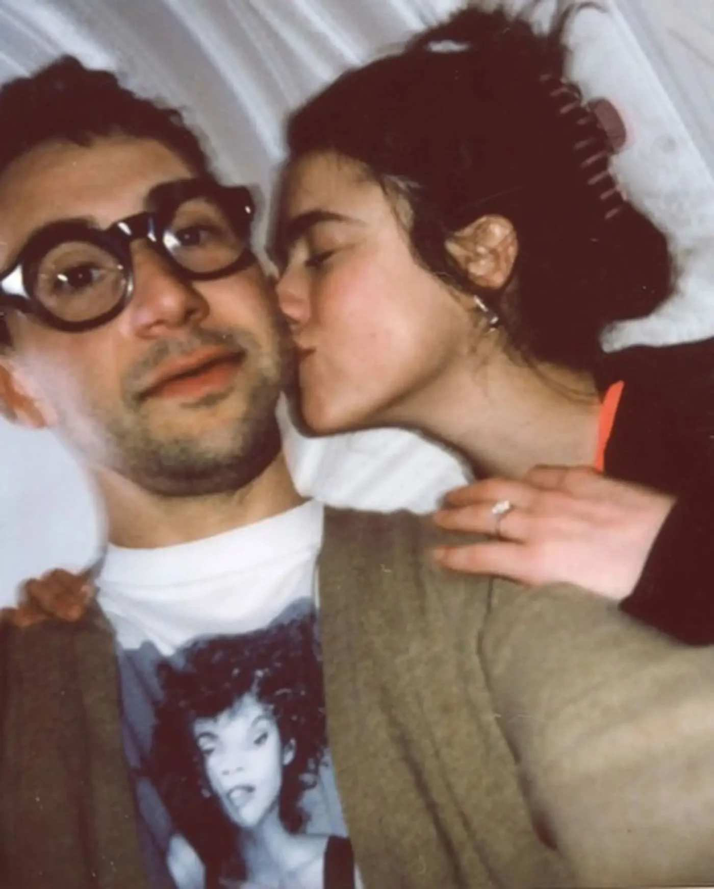 A person with dark hair and glasses looking at the camera, being kissed on the cheek by a woman with dark hair who is wearing a ring.