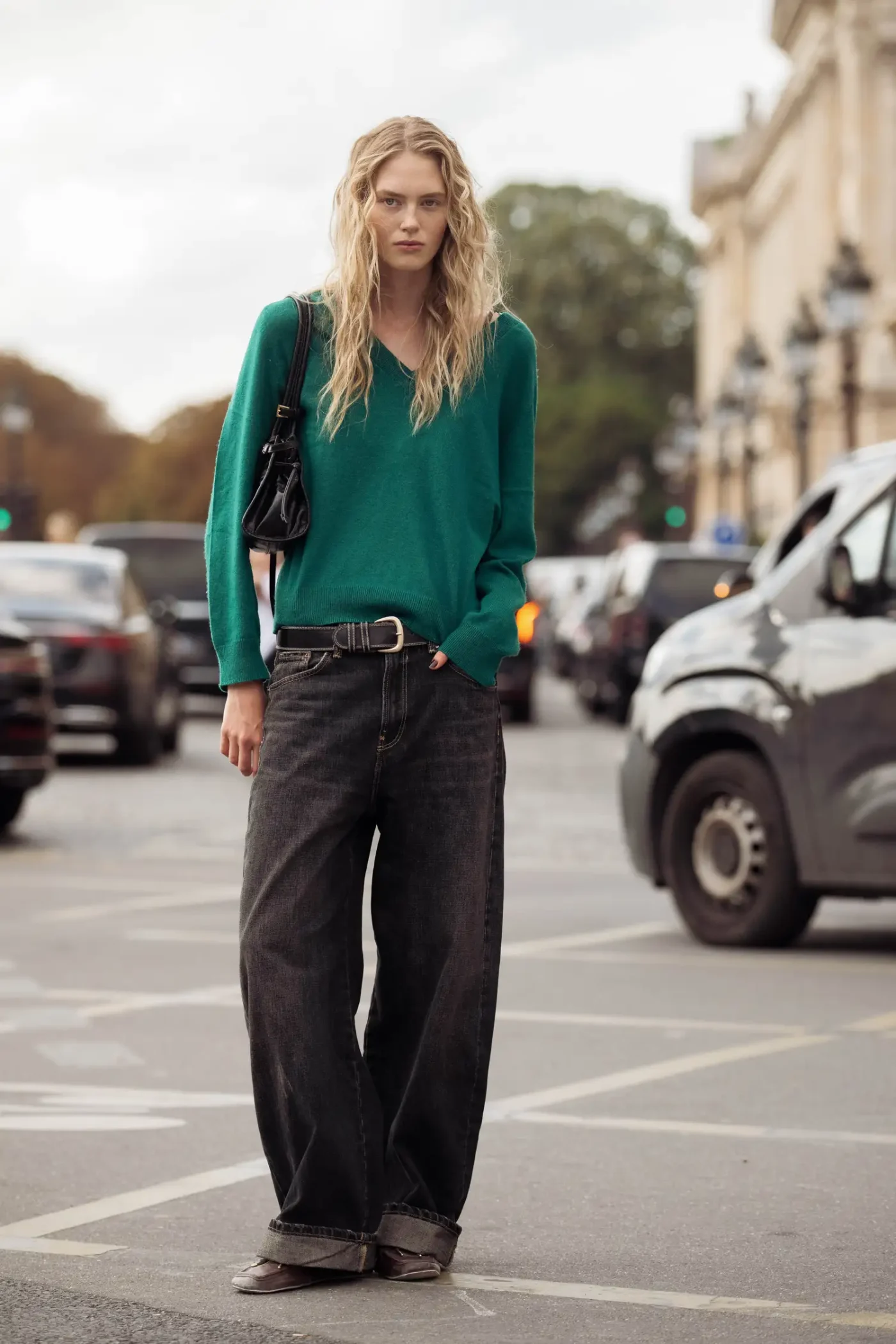A person with long hair wearing casual attire stands outdoors on a street near cars