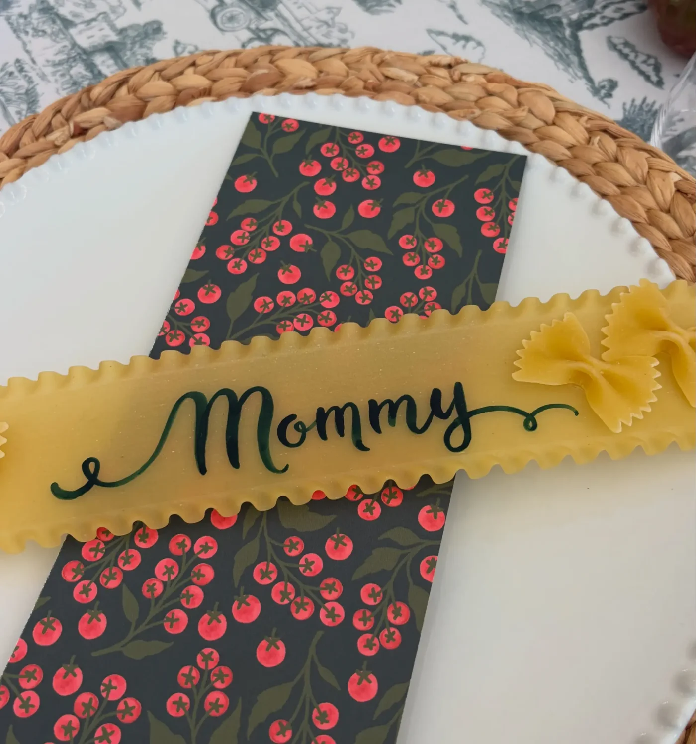 A place setting featuring a white plate, a patterned menu card, and a lasagna noodle with the word