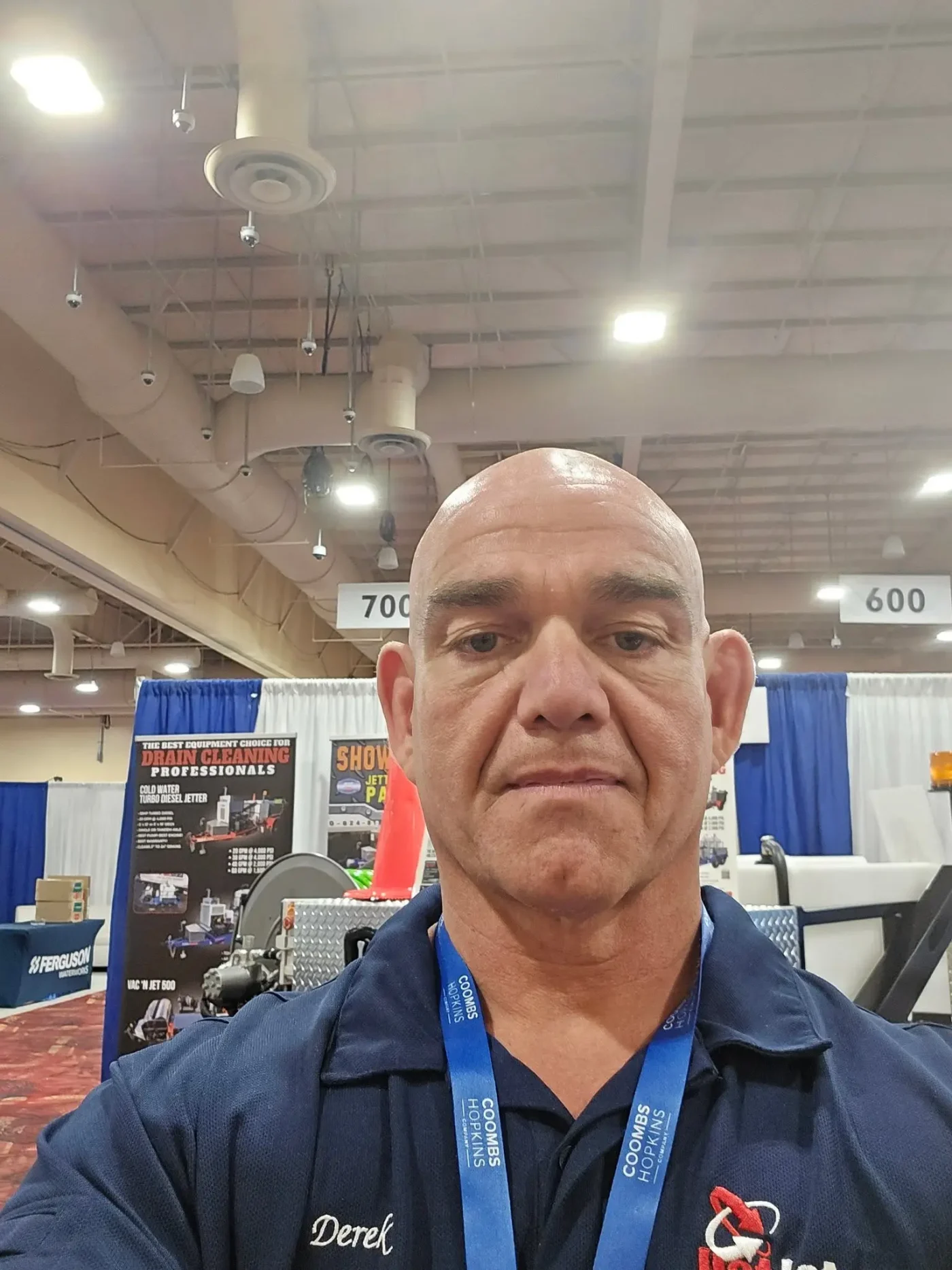 A selfie of Derek at an indoor exhibition hall, with trade show displays and hanging banners in the background.