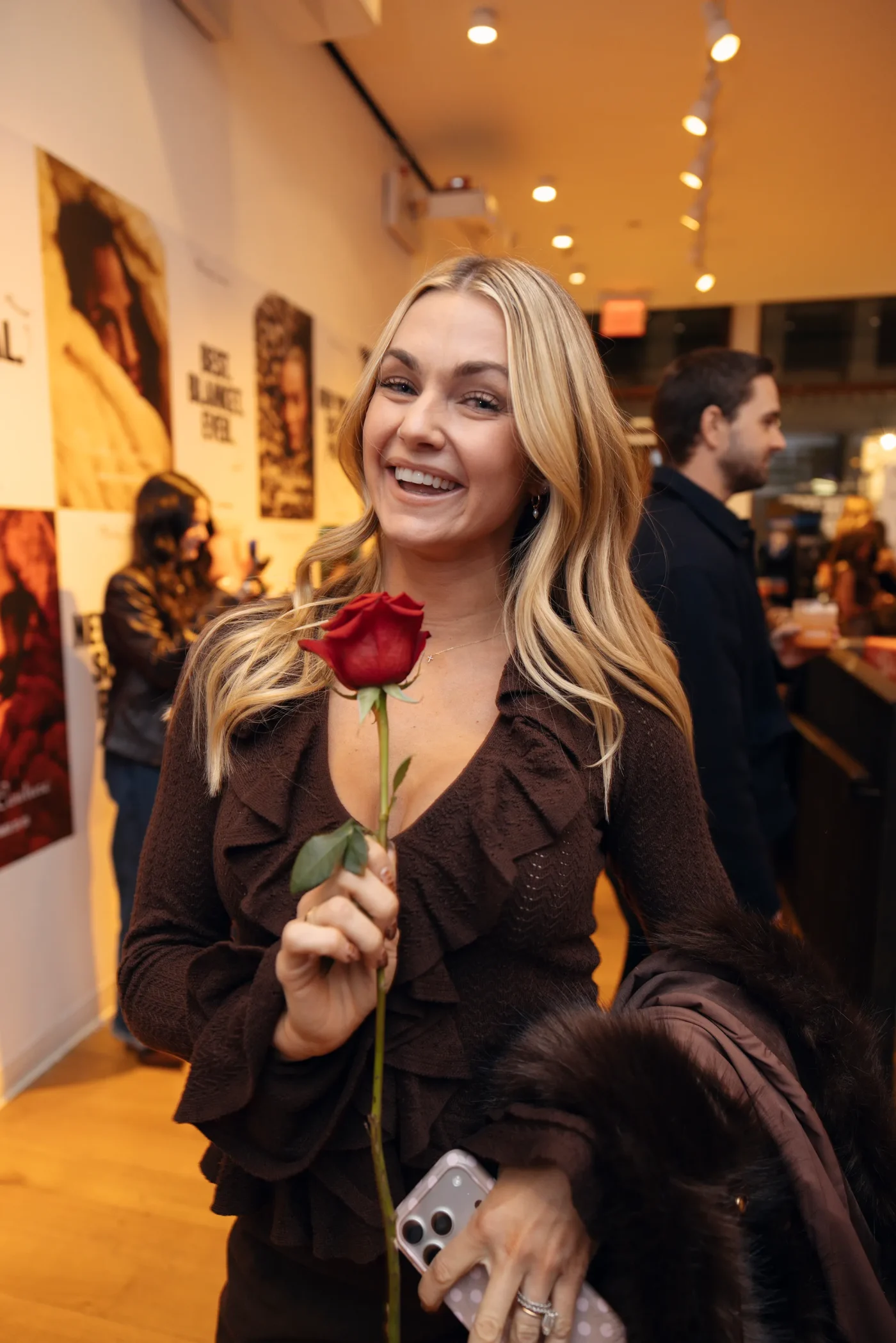 A smiling blond woman holds a red rose in an indoor setting.