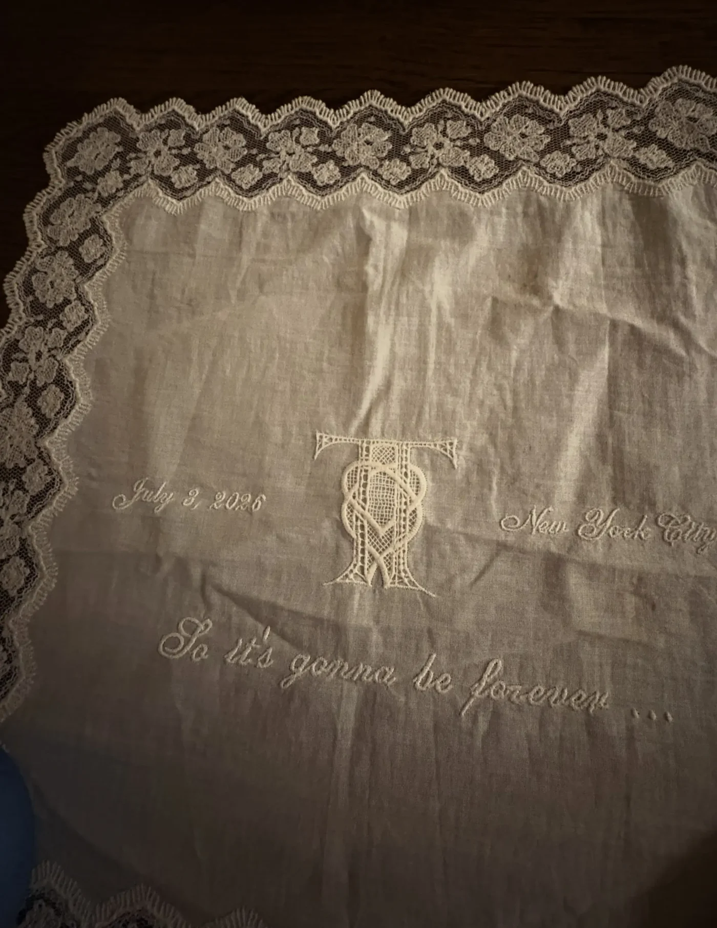 A white lace-trimmed wedding handkerchief embroidered with a monogram of the letters T and T, the location "New York City", and the lyric "So it
