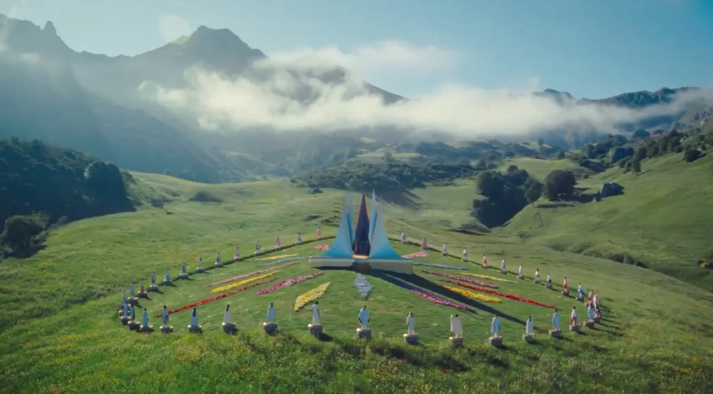 A wide shot of the arena in Hunger Games: Sunrise on the Reaping, showing tributes standing in a large circle on pedestals around a central structure, set in a lush green valley with mountains in the background.