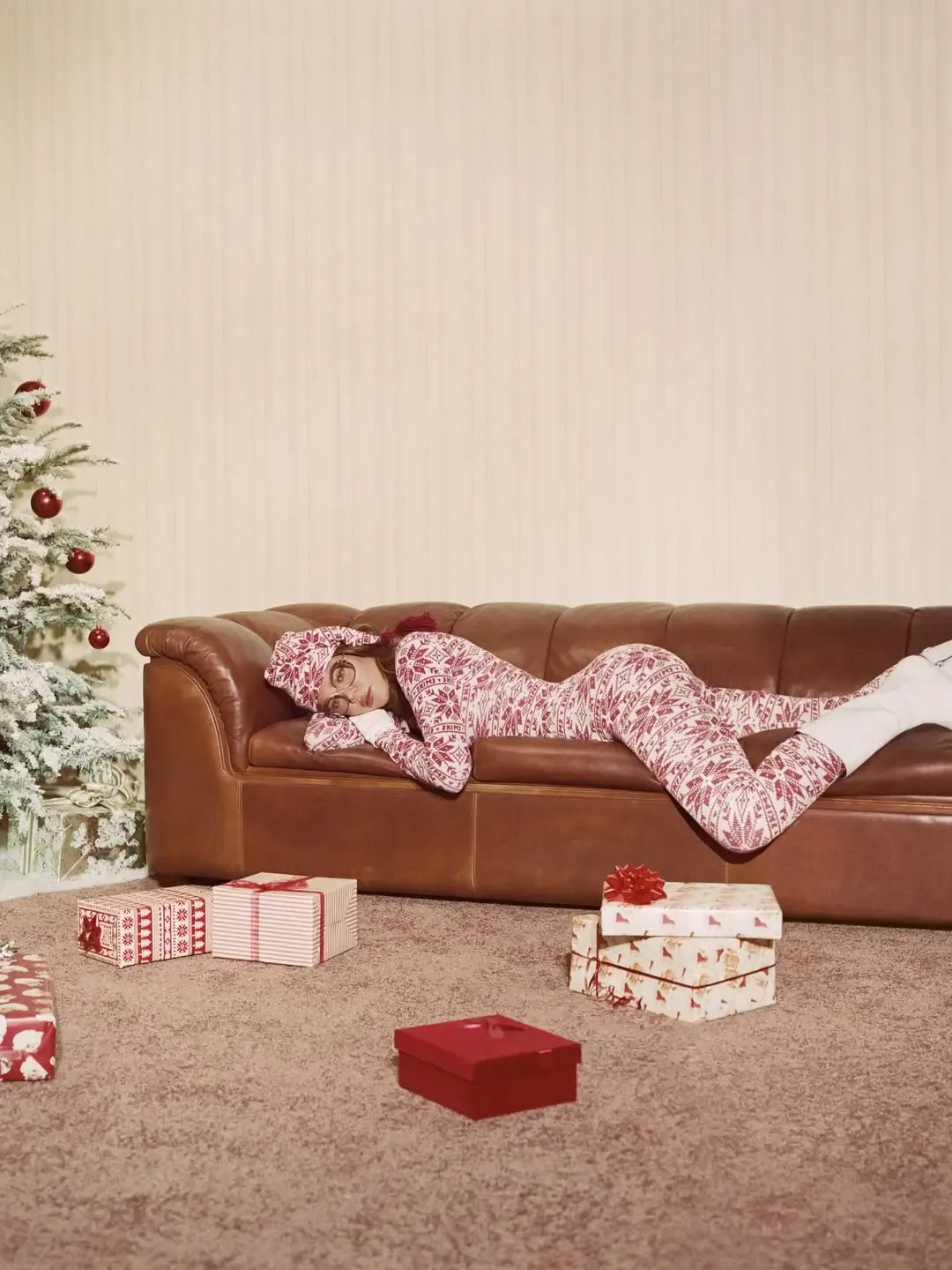 A woman in a matching red and white patterned Skims loungewear set rests on a brown leather couch in a room decorated for Christmas with a snowy tree and wrapped presents.