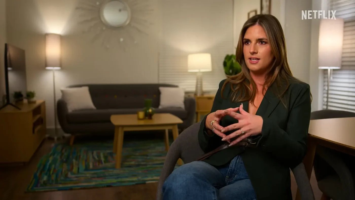 A woman sitting in a living room, speaking and gesturing with her hands, wearing a dark green jacket and blue jeans.
