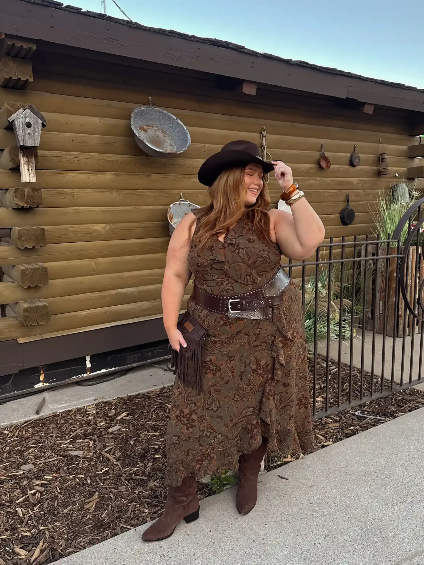 A woman wearing a cowboy hat standing in front of a rustic log cabin dressed in westernstyle clothing