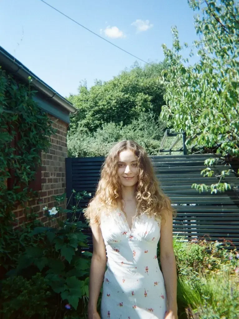 A woman with long blonde hair stands in a garden wearing a white floral dress.