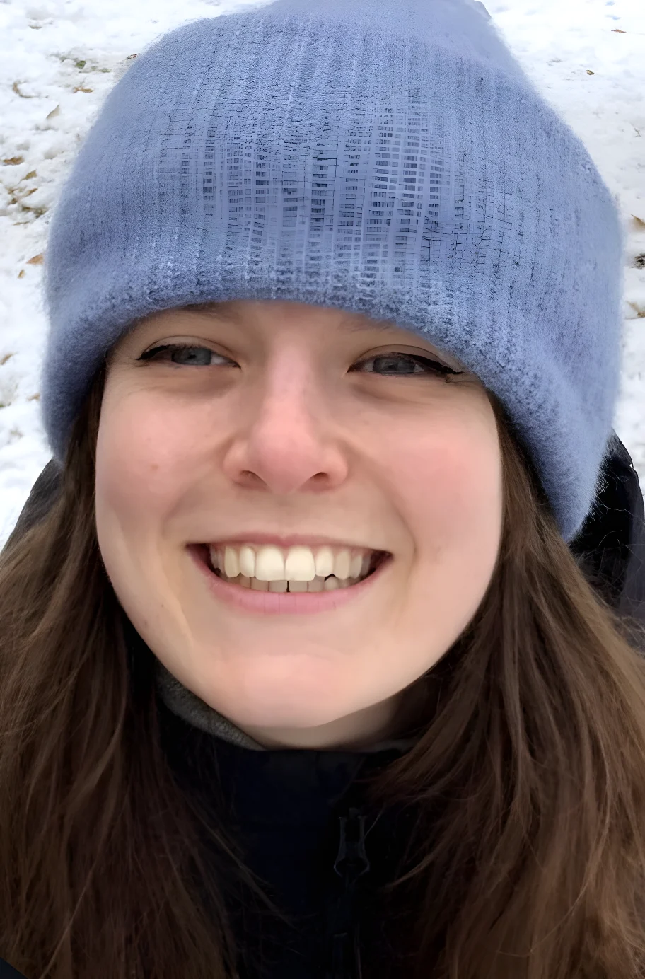 A woman with long brown hair smiles widely at the camera, wearing a grey knitted beanie and dark jacket, with snow visible in the background.