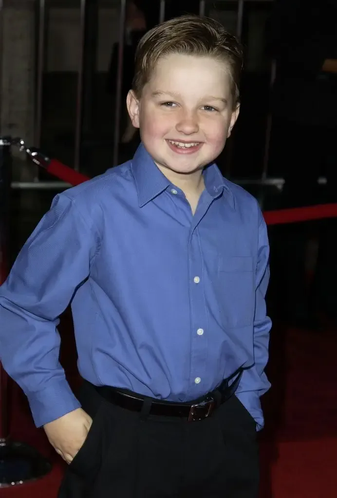 A young Angus T. Jones smiles broadly on the red carpet, wearing a blue button-down shirt and black trousers.