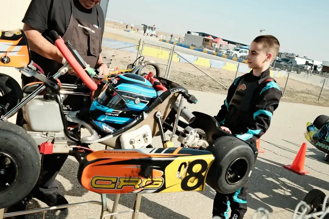 A young boy in racing gear stands beside a go-kart while an adult adjusts it.