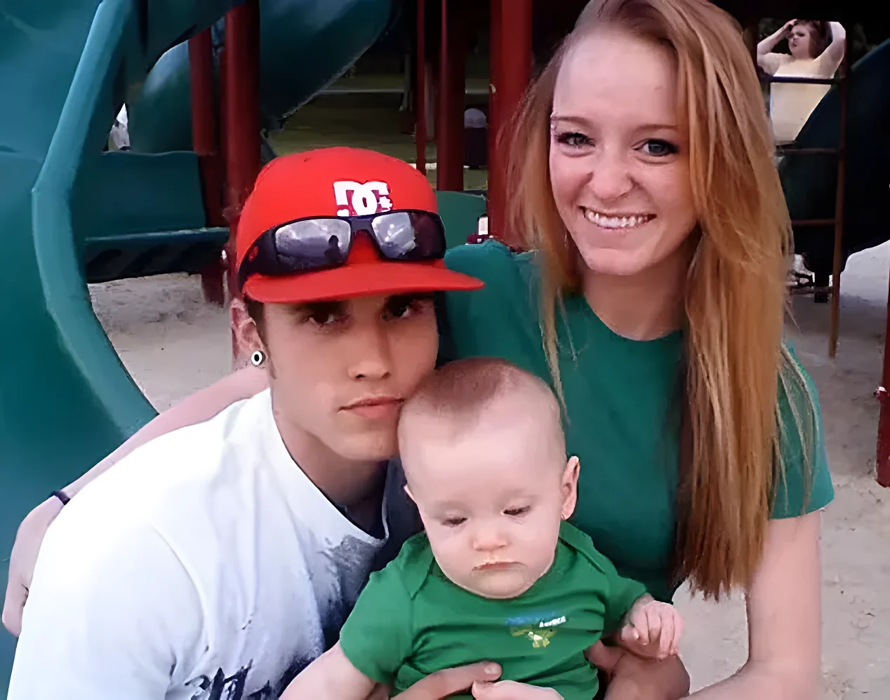 A young family of three poses together outdoors at a playground.