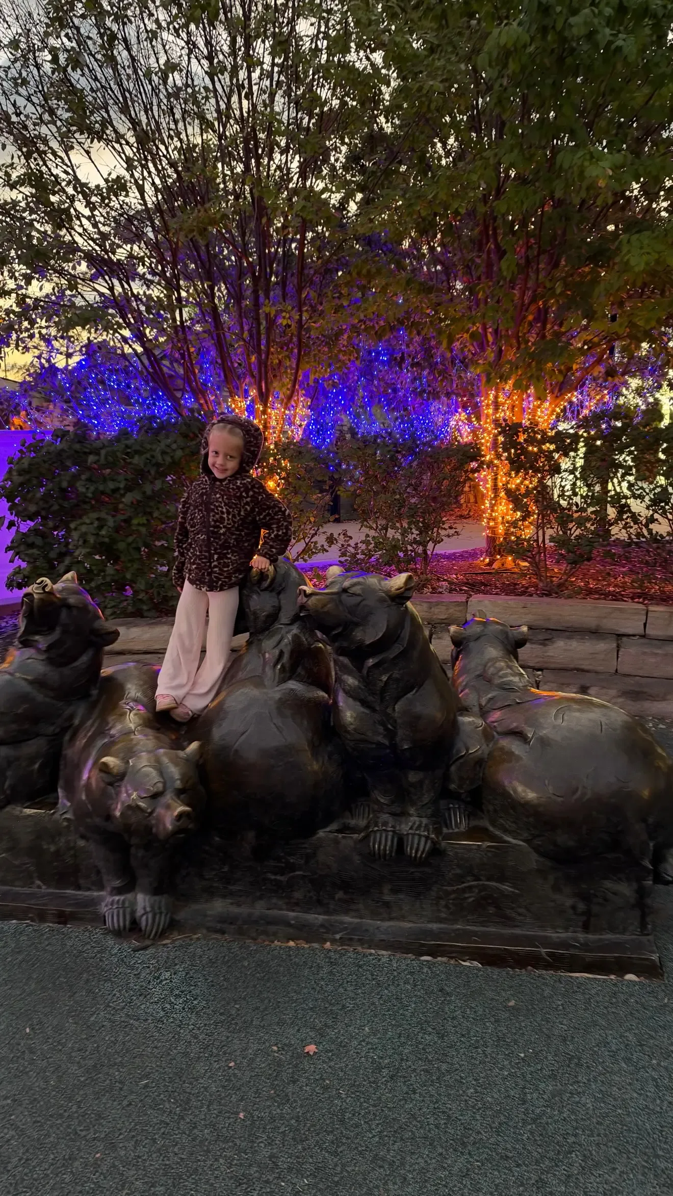 A young girl posing with bronze bear statues at night, with illuminated trees and colorful lights in the background.