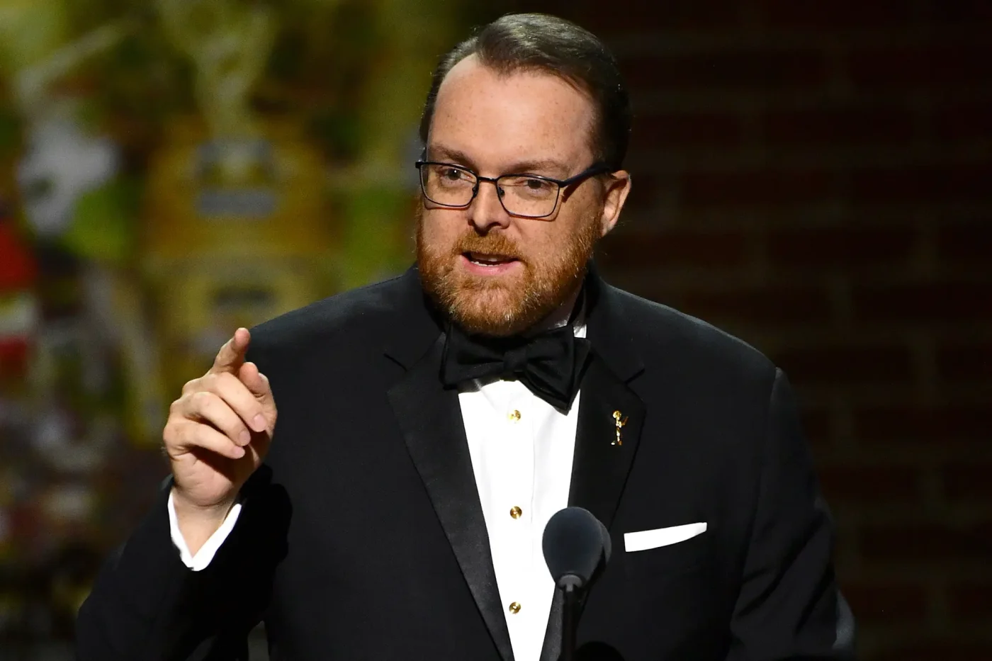 Actor and writer Dave Horwitz speaking at a podium while wearing a tuxedo and glasses.