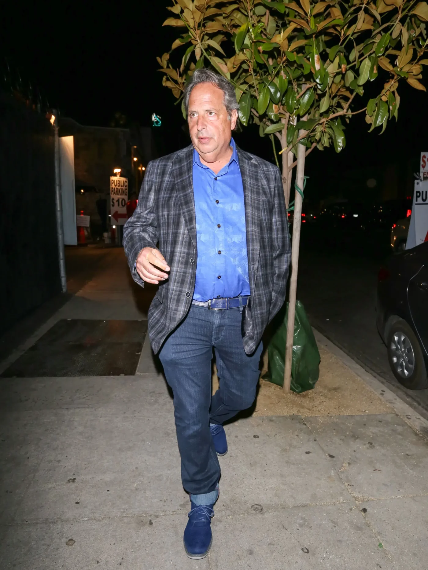 Actor Jon Lovitz walks along a sidewalk at night, wearing a plaid blazer, blue shirt, and jeans.