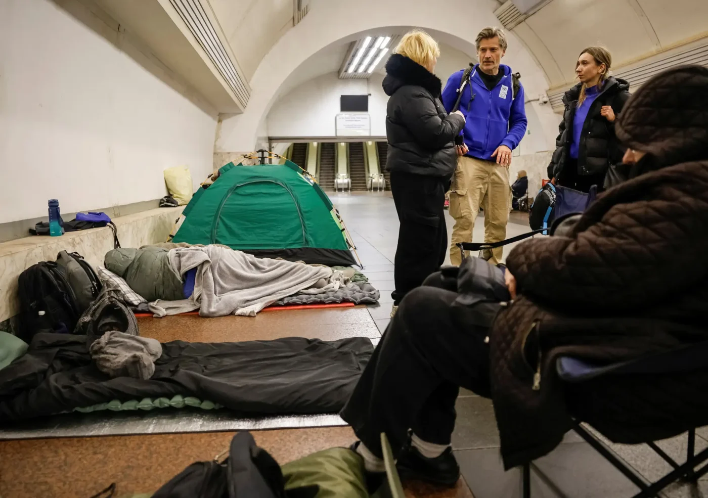 Actor Nikolaj Coster-Waldau in a blue hoodie stands talking to people inside a Kyiv metro station used as a shelter, with a green tent and sleeping people on the platform.