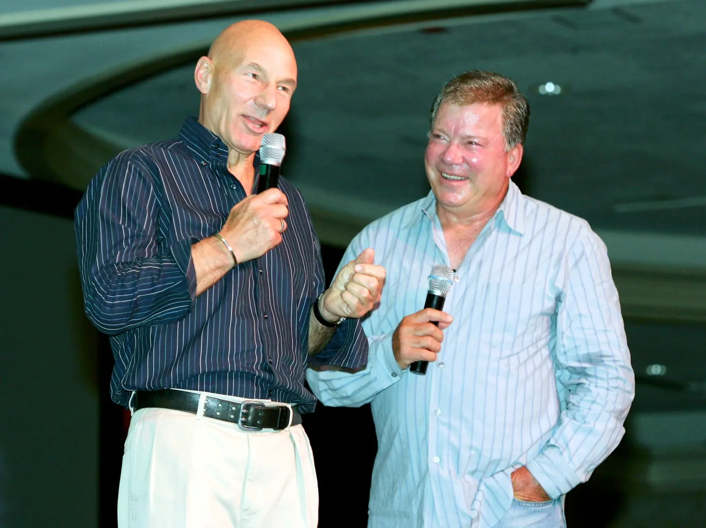 Actors Patrick Stewart and William Shatner holding microphones and speaking on stage at a Star Trek convention.