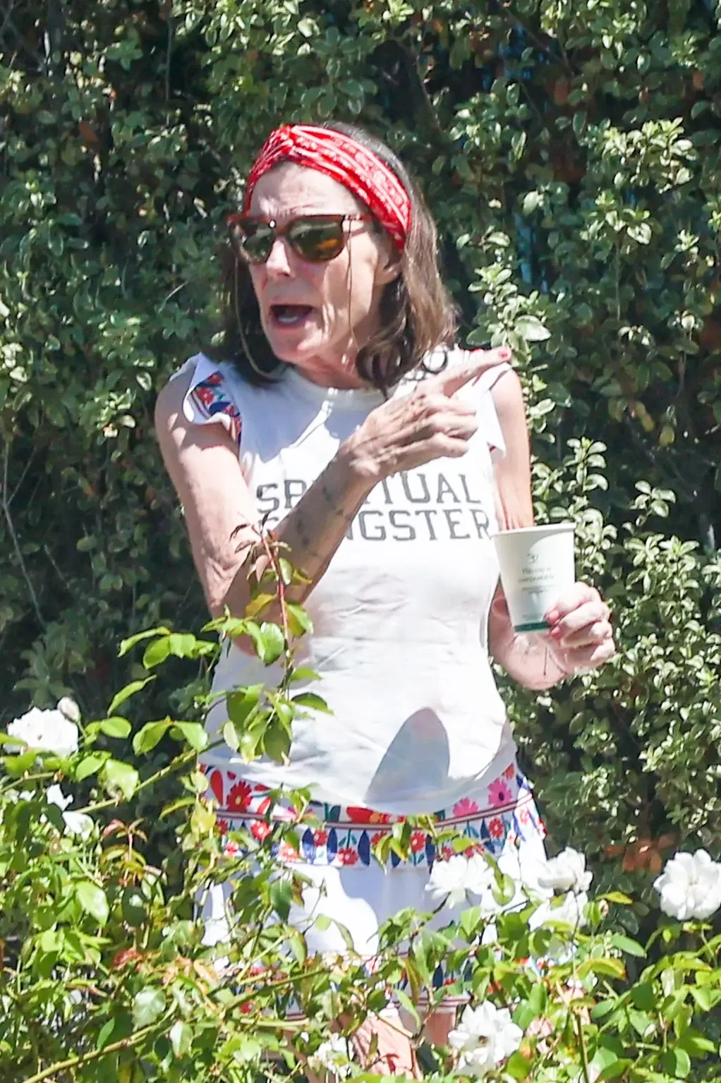Actress Susan St James wearing a red headband, sunglasses, and a white t-shirt while holding a cup outdoors near leafy bushes.
